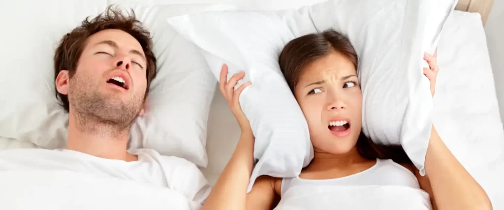a couple in bed, man is snoring but woman is awake and covering her ears